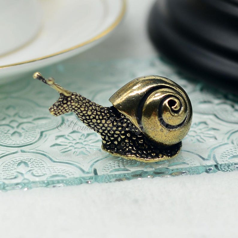 Puede incluir: Una peque&ntilde;a figurita de caracol de color dorado con una concha y un cuerpo detallados. El caracol est&aacute; sentado sobre una superficie de vidrio transparente con un patr&oacute;n decorativo.