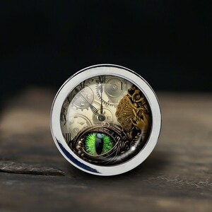 Peut inclure: Un cadran d'horloge argenté avec des chiffres romains et un œil vert au centre. Le cadran est entouré d'engrenages et d'autres éléments d'inspiration steampunk.