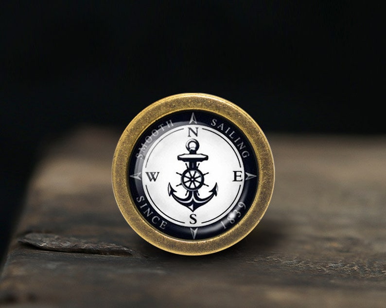 May include: A gold-coloured compass with a black and white anchor design. The compass face reads "Smooth Sailing Since 1839" and has the cardinal directions N, S, E, and W.