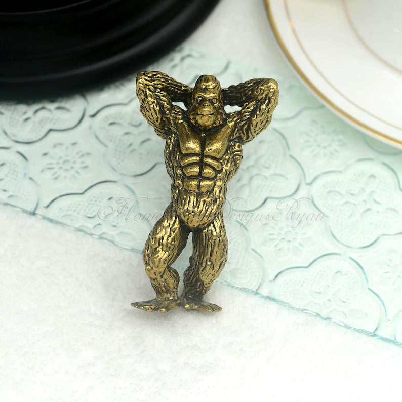 Op de afbeelding: Een kleine, goudkleurige gorilla-figuur met de armen achter het hoofd. Het beeldje heeft gedetailleerde vacht en spierdefinitie en staat op een lichtblauwe ondergrond. De gorilla is een decoratief item.