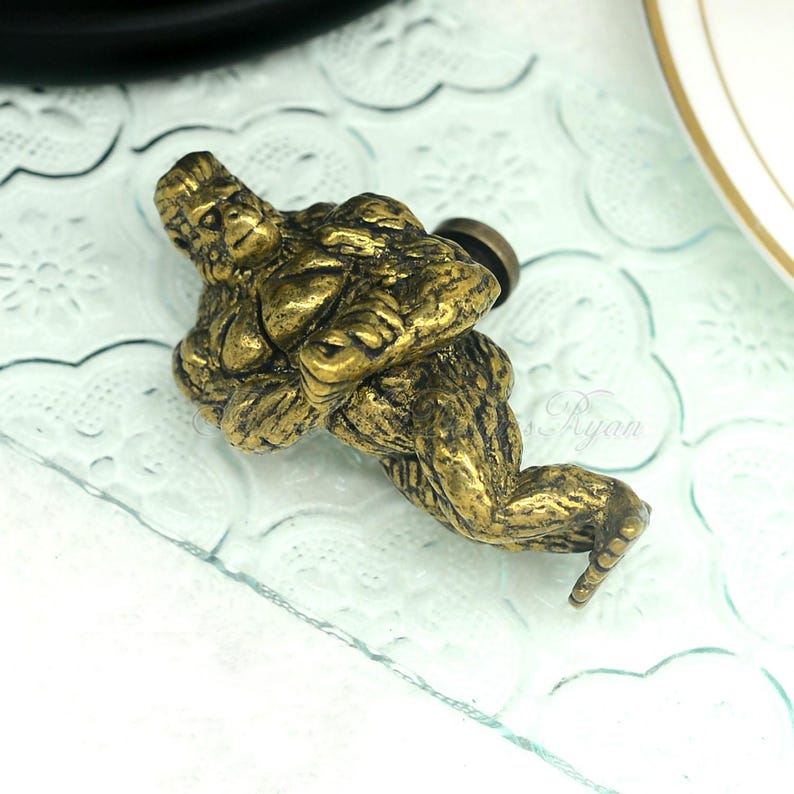 Puede incluir: Un pomo de armario de metal dorado con forma de mono agachado. El mono tiene una superficie detallada y texturizada.