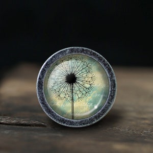 Op de afbeelding: Een ronde, zilverkleurige, decoratieve knop met een glazen inzetstuk met een blauwe en groene aquarel afbeelding van een paardenbloem.