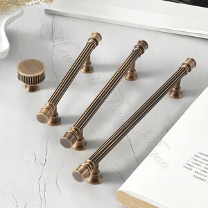 Puede incluir: Un juego de herrajes de gabinete de bronce antiguo, que incluye tres tiradores largos y un pomo redondo. Los tiradores tienen un dise&ntilde;o acanalado y son adecuados para cajones o puertas. Los herrajes se exhiben sobre una superficie clara.