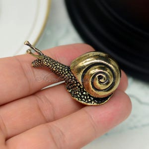 Puede incluir: Una figurita de caracol de color dorado con una concha detallada y un cuerpo texturizado. El caracol mide aproximadamente 5 cm de largo y est&aacute; hecho de metal.