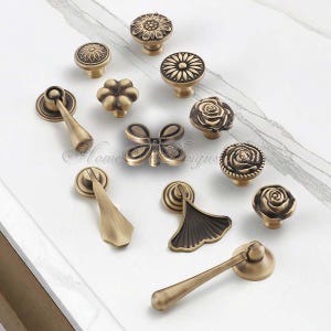 Pu&ograve; includere: Una collezione di pomelli e maniglie per mobili in ottone antico con vari disegni floreali e geometrici. L'hardware include pomelli a forma di fiori, rose e raggi di sole, oltre a maniglie a farfalla e a ventaglio.