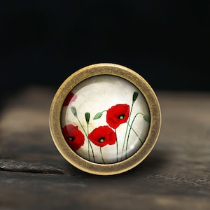Peut inclure: Un pendentif rond de couleur laiton avec un dôme en verre représentant une illustration de style vintage de coquelicots rouges sur un fond crème.