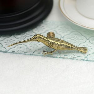 May include: Gold-toned metal hummingbird drawer pull with a detailed design. The pull is shown on a light blue surface.