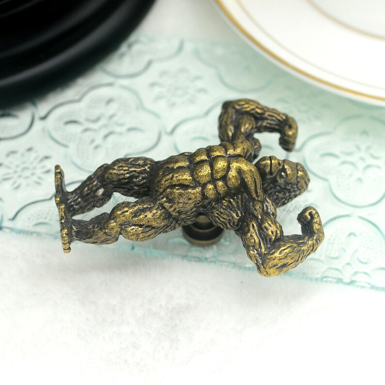 Pu&ograve; includere: Un pomello per armadio in metallo dorato a forma di gorilla con le braccia flesse. Il gorilla &egrave; in posizione accovacciata.