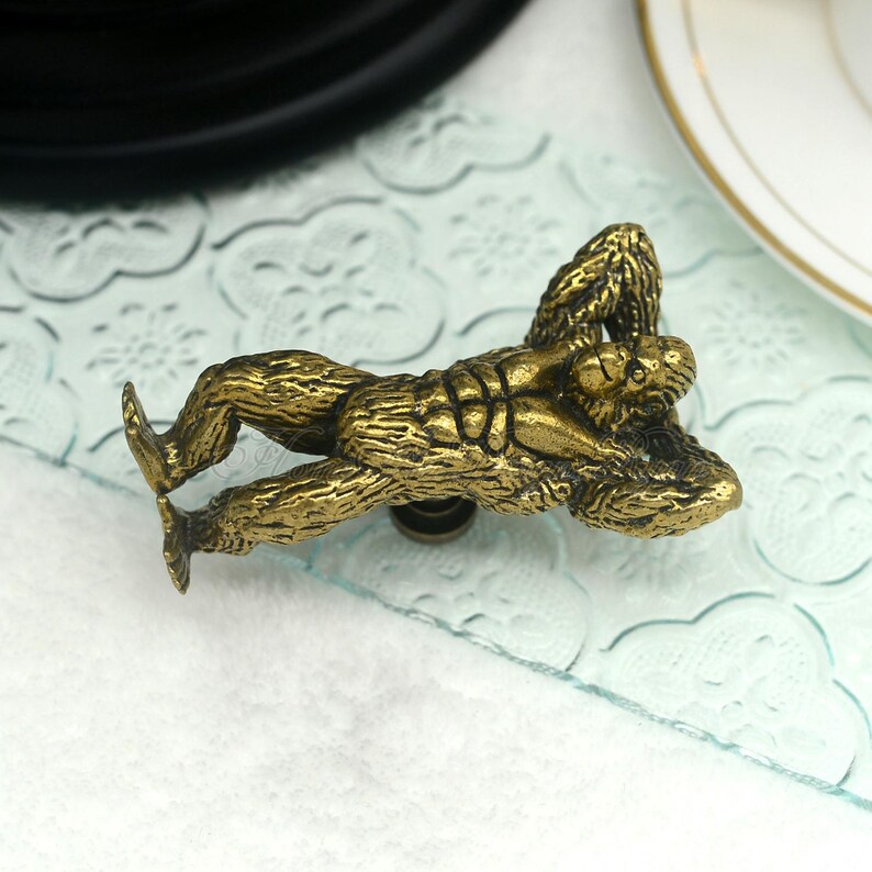 Op de afbeelding: Een bronzen kastknop in de vorm van een liggende gorilla. De gedetailleerde hardware heeft een getextureerde afwerking en is gemonteerd op een helder glazen oppervlak. De gorilla ligt met uitgestrekte armen en benen.