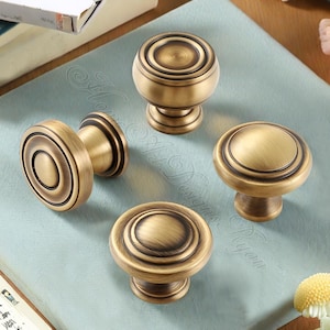 May include: A set of antique brass cabinet knobs. The knobs feature a circular design with concentric rings and a warm, aged finish. The hardware is likely for furniture or decorative purposes.
