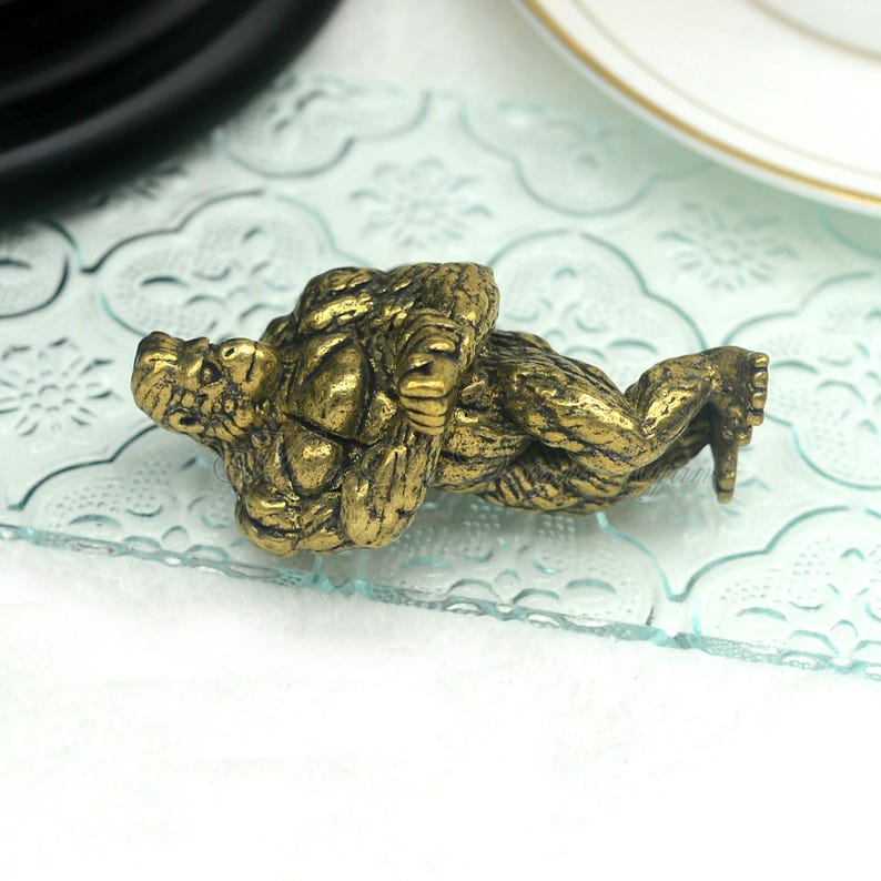 Puede incluir: Una figurita de metal de color dorado de un gorila acostado de espaldas con los brazos cruzados sobre el pecho.