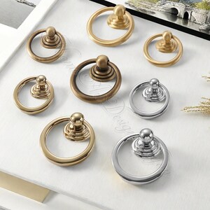 Puede incluir: Una variedad de tiradores de armario en acabados dorados, bronce y plateados. Cada tirador presenta un asa circular unida a una base decorativa. El hardware est&aacute; dispuesto sobre una superficie blanca, mostrando los diferentes estilos. Medidas en cent&iacute;metros.