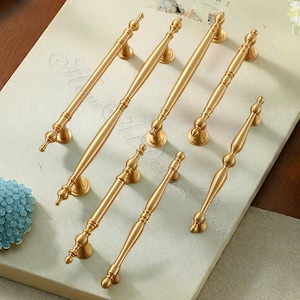 Puede incluir: Siete tiradores dorados para armarios con detalles decorativos. Los tiradores est&aacute;n dispuestos sobre una superficie de color claro, mostrando su elegante dise&ntilde;o. Cada tirador presenta un agarre central y extremos ornamentados.