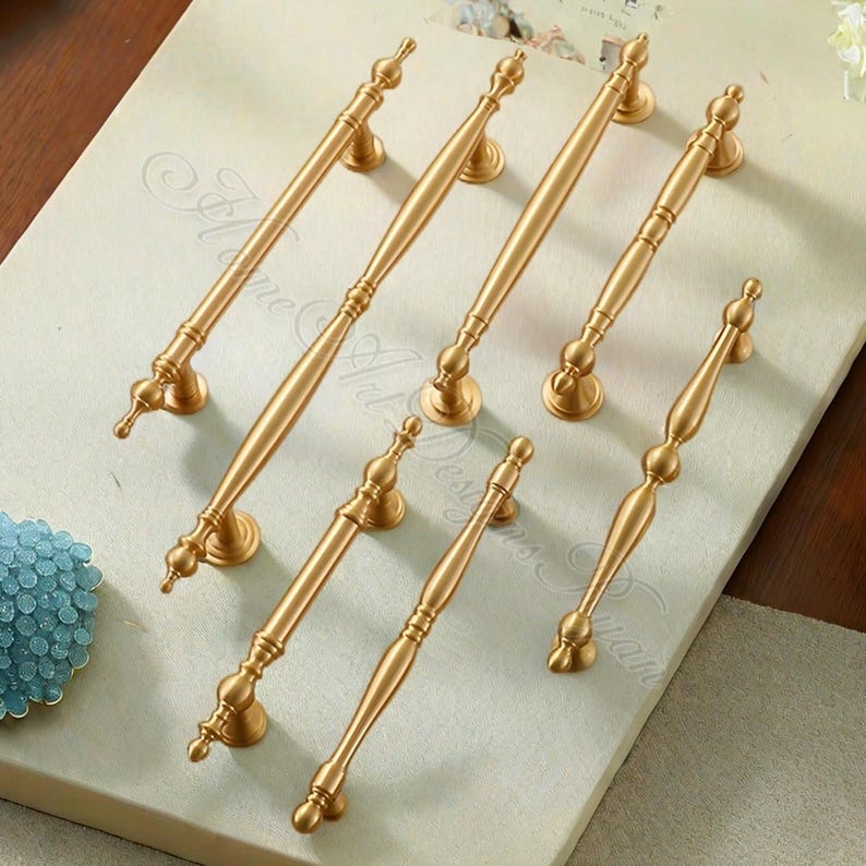 Puede incluir: Siete tiradores dorados para armarios con detalles decorativos. Los tiradores est&aacute;n dispuestos sobre una superficie de color claro, mostrando su elegante dise&ntilde;o. Cada tirador presenta un agarre central y extremos ornamentados.