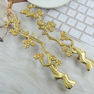 May include: Two gold-toned cabinet pulls shaped like stylized branches with blossoms. The pulls are on a white surface.