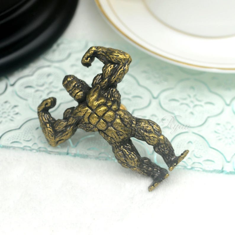 Pu&ograve; includere: Un pomello per armadio in metallo dorato a forma di creatura scimmiesca muscolosa con le braccia alzate in una posa di flessione.