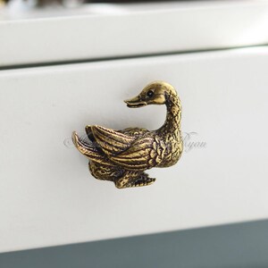 Op de afbeelding: Een gouden eendenvormige ladegreep. De eend kijkt naar rechts en heeft zijn vleugels uitgespreid.