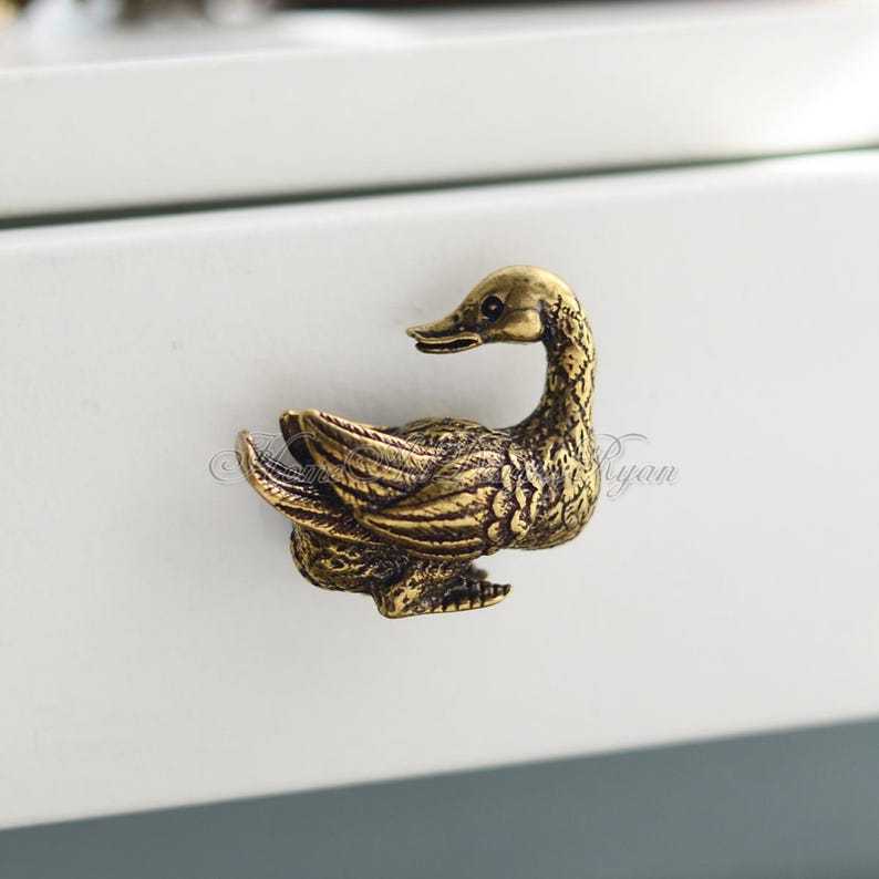 Op de afbeelding: Een gouden eendenvormige ladegreep. De eend kijkt naar rechts en heeft zijn vleugels uitgespreid.