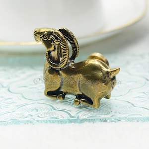 May include: A gold-toned metal ram figurine with detailed horns and a standing pose. The ram is facing to the right and has a textured surface.