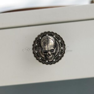 May include: A silver skull drawer pull with an intricate design. The skull is centrally positioned within a decorative, scalloped border. The hardware is mounted on a white surface.