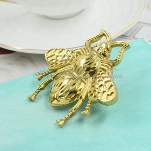 Puede incluir: Un broche de abeja de color dorado con detalles intrincados. La abeja tiene un cuerpo y alas texturizados.