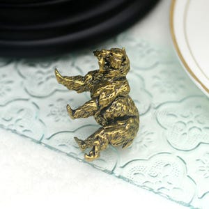 May include: A gold-colored metal bear figurine sitting on a light blue patterned surface. The bear is sitting up with its paws raised and its head tilted back.