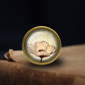 May include: A gold-toned round metal button with a glass top featuring a vintage illustration of a white poppy flower with a brown center.