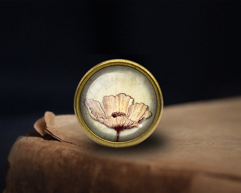 May include: A gold-toned round metal button with a glass top featuring a vintage illustration of a white poppy flower with a brown center.
