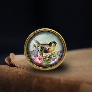 Peut inclure: Un bouton rond en verre recouvert d'or avec une illustration vintage d'un petit oiseau perché sur une branche avec des fleurs roses et violettes.