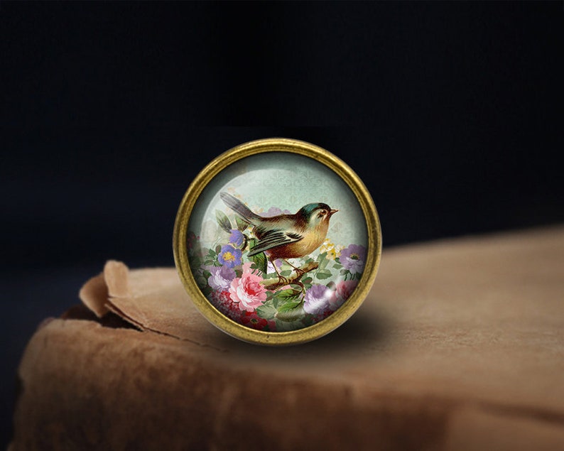 K&ouml;nnte beinhalten: Ein runder, goldfarbener, glas&uuml;berzogener Knopf mit einer Vintage-Illustration eines kleinen Vogels, der auf einem Zweig mit rosa und violetten Blumen sitzt.