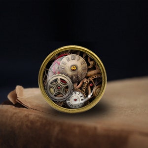 Peut inclure: Un pendentif rond en verre recouvert d'or avec un design steampunk comprenant des engrenages, des cadrans d'horloge et des chiffres romains. Le pendentif a un bijou rouge au centre d'un des engrenages.
