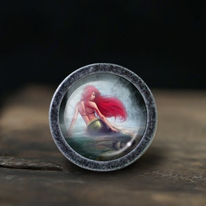 Puede incluir: Perilla de armario redonda, plateada, con una sirena de largo cabello rojo. La sirena está representada sentada, con una cola verde y un top azul y dorado. La perilla tiene un borde texturizado de estilo antiguo.