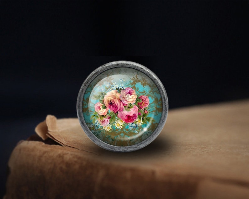 May include: A round, silver-toned cabinet knob with a glass insert featuring a vintage floral design. The design includes pink, white, and yellow roses on a blue and gold background.