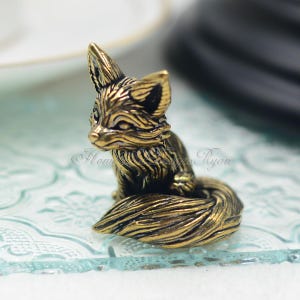 Puede incluir: Una pequeña figurita de metal dorado de un zorro sentado con la cola enroscada alrededor de su cuerpo. El zorro tiene una superficie detallada, texturizada y una apariencia realista.