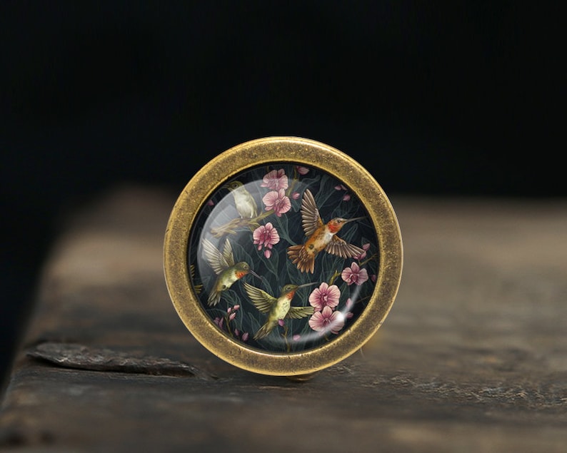May include: A round brass knob with a glass insert featuring a floral design with hummingbirds in shades of pink, green, and orange.