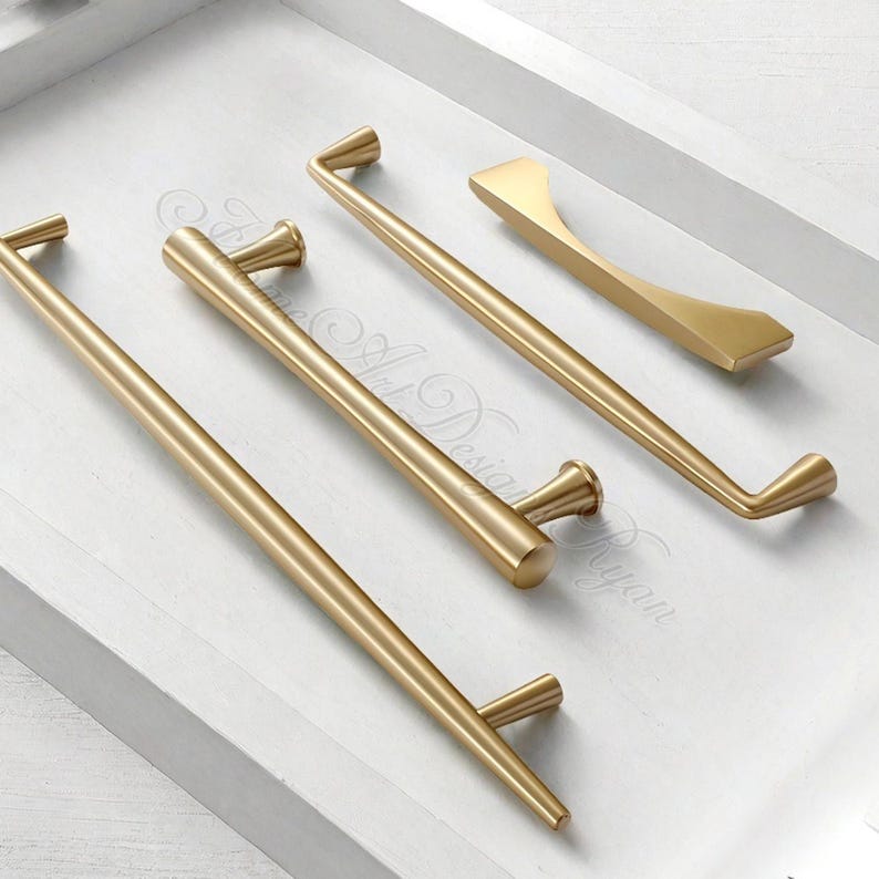 Puede incluir: Un conjunto de herrajes dorados para armarios, que incluyen tiradores de barra y un tirador, dispuestos sobre una superficie blanca. Los herrajes presentan varios dise&ntilde;os, con formas redondeadas y angulares. La est&eacute;tica general es moderna y minimalista.