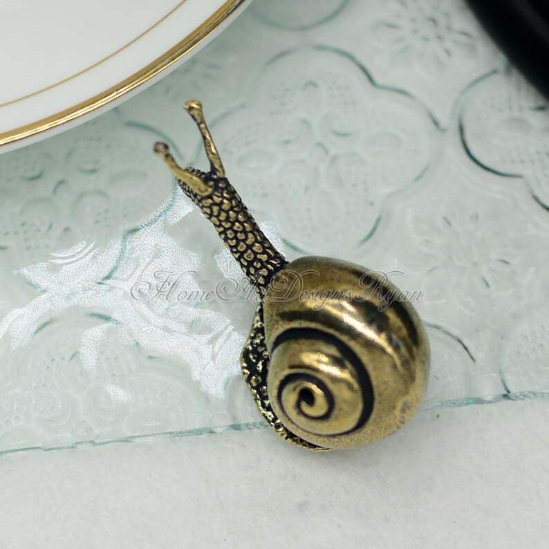 Puede incluir: Una figurita de caracol de metal de color dorado con una concha detallada y un cuerpo texturizado. El caracol est&aacute; sentado sobre una superficie de vidrio con un patr&oacute;n floral.