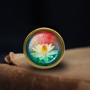 May include: A round, brass-colored pendant with a glass dome featuring a colorful watercolor painting of a white lotus flower with a yellow center. The background is a mix of pink, blue, and green.