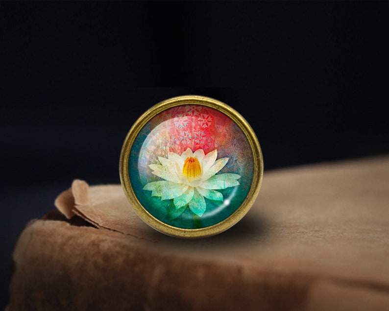 May include: A round, brass-colored pendant with a glass dome featuring a colorful watercolor painting of a white lotus flower with a yellow center. The background is a mix of pink, blue, and green.