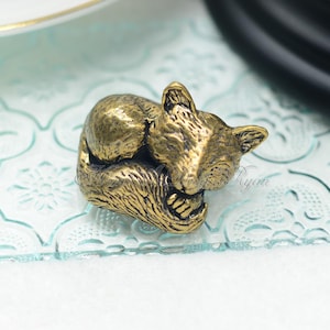 May include: A detailed, antique-style brass figurine of a sleeping cat. The cat is curled up, with its paws tucked in and its head resting on its body. The figurine has a textured surface and is finished in a warm, golden hue. The cat figurine is a decorative item.