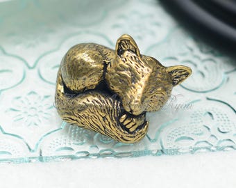 May include: A detailed, antique-style brass figurine of a sleeping cat. The cat is curled up, with its paws tucked in and its head resting on its body. The figurine has a textured surface and is finished in a warm, golden hue. The cat figurine is a decorative item.