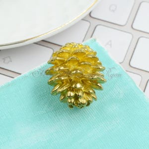 May include: A gold-colored metal pine cone ornament. The ornament is detailed and realistic.