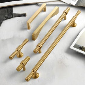 Puede incluir: Una colecci&oacute;n de herrajes dorados para armarios, que incluye varios dise&ntilde;os de tiradores y pomos. Los tiradores var&iacute;an en longitud y forma, algunos con un dise&ntilde;o de barra recta y otros con elementos curvos o angulados. Los pomos tienen forma cil&iacute;ndrica con base circular.