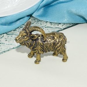 May include: A small, gold-colored metal figurine of a goat with curled horns. The goat is standing on a white surface.