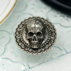 May include: A silver-coloured skull-shaped decorative knob. The skull features a crack down the middle and is set within a decorative, scalloped edge. The knob is likely made of metal and is designed for furniture or decor.