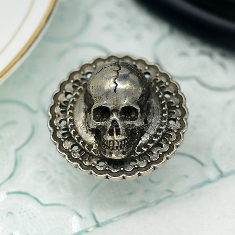 May include: A silver-coloured skull-shaped decorative knob. The skull features a crack down the middle and is set within a decorative, scalloped edge. The knob is likely made of metal and is designed for furniture or decor.