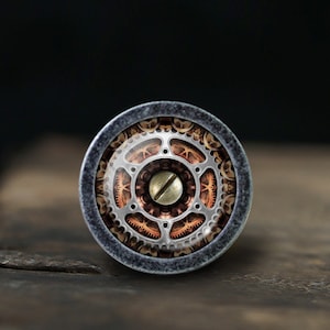 Peut inclure: Un bouton rond en argent avec un motif détaillé d'engrenages et de pignons dans des tons de cuivre et d'argent. Le bouton a une petite vis en argent au centre.