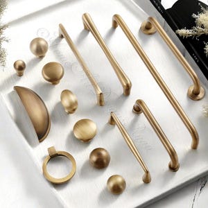 Può includere: Una collezione di ferramenta per mobili in ottone antico, tra cui pomelli, maniglie e un anello. L'hardware presenta una calda tonalità dorata e varie forme, disposte su una superficie bianca. Diversi design per l'arredamento della casa.