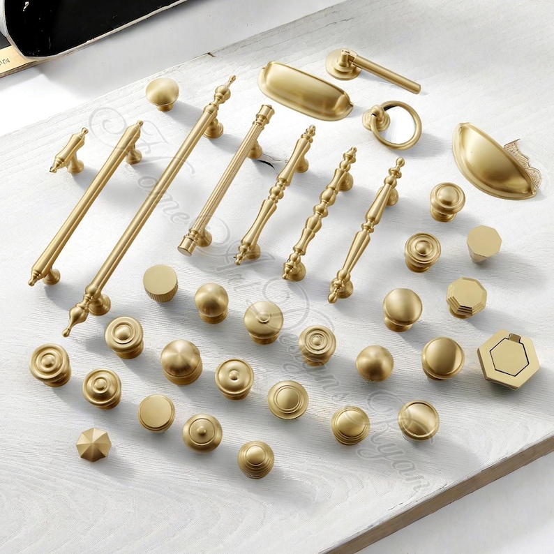 Puede incluir: Una colecci&oacute;n de herrajes dorados para armarios, que incluye varios pomos, tiradores y manijas. Los herrajes est&aacute;n dispuestos sobre una superficie de madera blanca, mostrando diferentes dise&ntilde;os y tama&ntilde;os. Los herrajes son de metal.