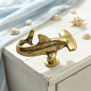 May include: A brass-toned metal cabinet knob shaped like a hammerhead shark. The shark figure is depicted in a swimming position with its tail curved upward and its fins extended. The knob is mounted on a small, circular, flat-topped base.